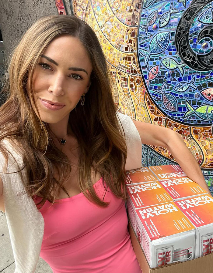 Woman in a pink top holding a box of "Holy Water" against a mosaic wall, relating to American Pie star. Woman in a pink top holding a box of "Holy Water" against a mosaic wall, relating to American Pie star.