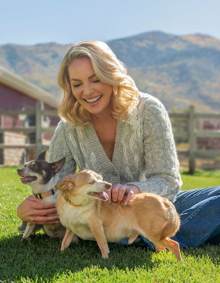 A woman joyfully playing with two small dogs on grass, surrounded by mountains. A woman joyfully playing with two small dogs on grass, surrounded by mountains.