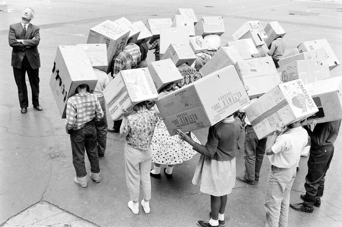 Children with cardboard boxes on their heads and a man observing, depicting life during JFK years in America.