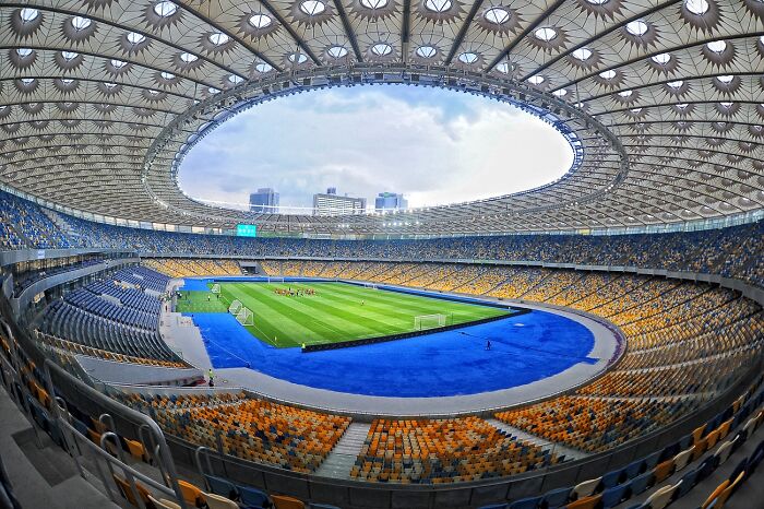Panoramic view of a large soccer cathedral stadium with vibrant seating and a green pitch under a patterned roof.