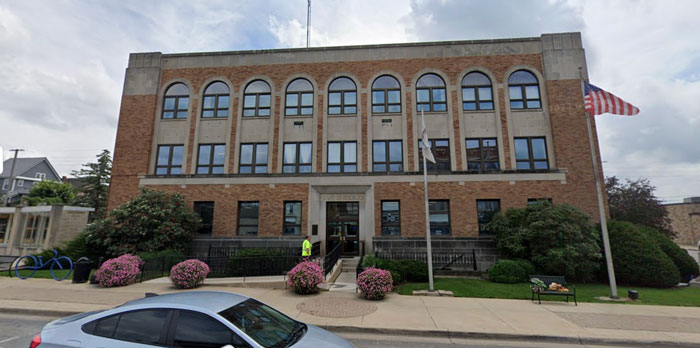 Street view of a police office building with flags, shrubs, and a parked car in front. Street view of a police office building with flags, shrubs, and a parked car in front.