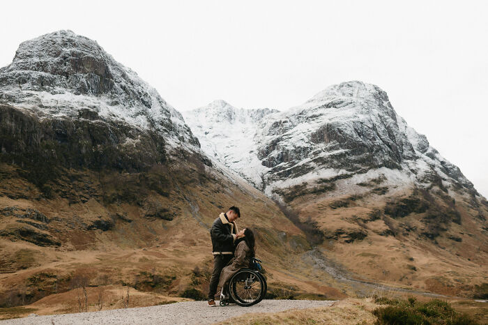 Couple in an engagement photo set against a stunning mountainous backdrop, with the woman seated in a wheelchair.