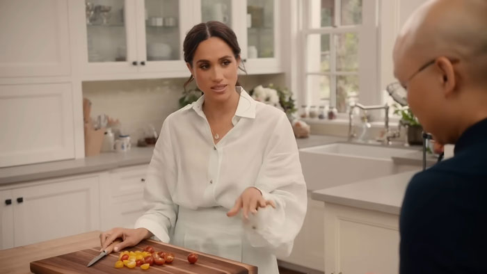Meghan Markle in a kitchen, discussing with a crew member, tomato slices on a cutting board, wearing a white blouse. Meghan Markle in a kitchen, discussing with a crew member, tomato slices on a cutting board, wearing a white blouse.
