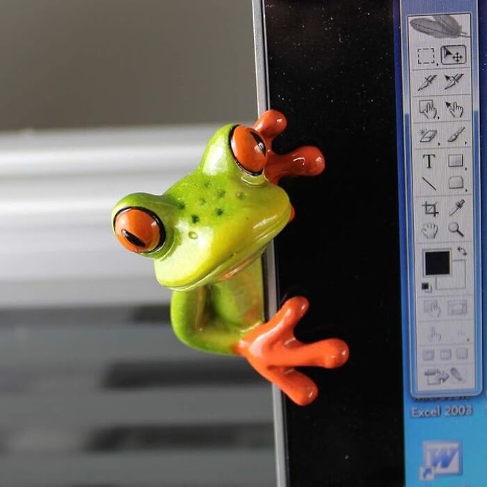 A small green and orange frog figurine clings to a computer monitor in an office, adding fun to the workday.