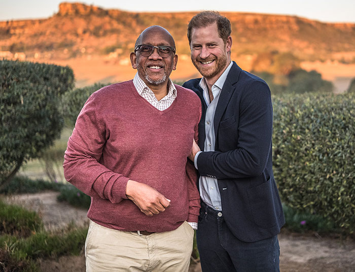 Two men smiling outdoors with greenery and sunset, related to Prince Harry and charity. Two men smiling outdoors with greenery and sunset, related to Prince Harry and charity.