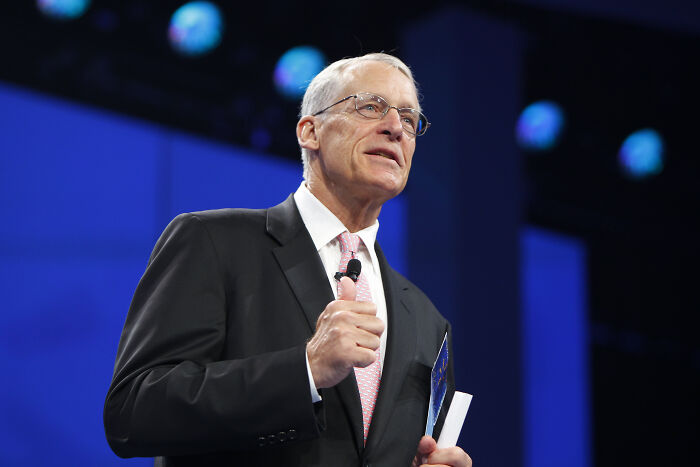 Older man in suit and glasses speaking on stage, symbolizing the richest people in the world and their wealth impact.