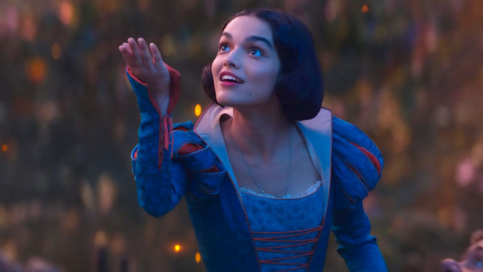 Actress in a blue dress as Snow White, smiling and reaching out against a forest backdrop, representing Disney's Snow White. Actress in a blue dress as Snow White, smiling and reaching out against a forest backdrop, representing Disney's Snow White.