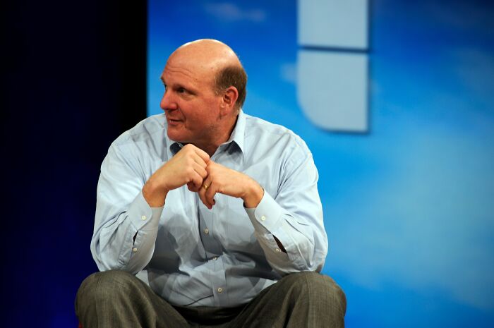 Bald man in light blue shirt sitting with hands clasped, representing one of the richest people in the world with vast wealth.