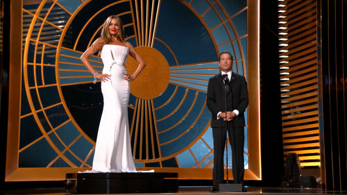 Two presenters on stage at an awards show, one in a striking white gown, the other in a black suit, amidst a dramatic backdrop.