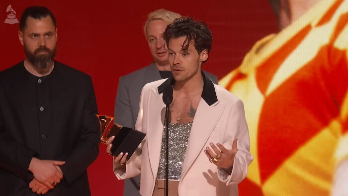 An artist in a white jacket holds an award, delivering a speech during a scandalous awards show moment.