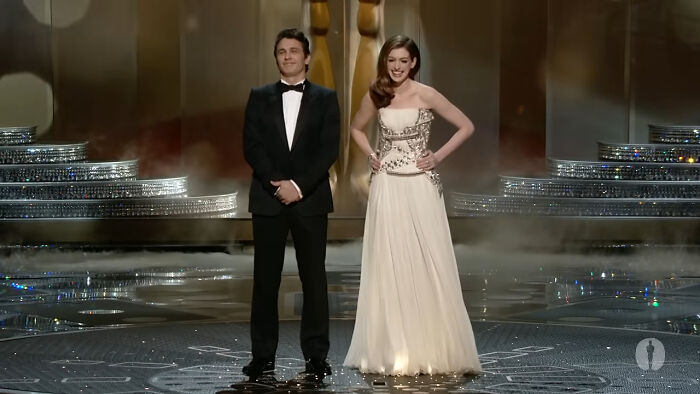 Hosts in formal attire presenting on a glittering stage during a memorable awards show moment.