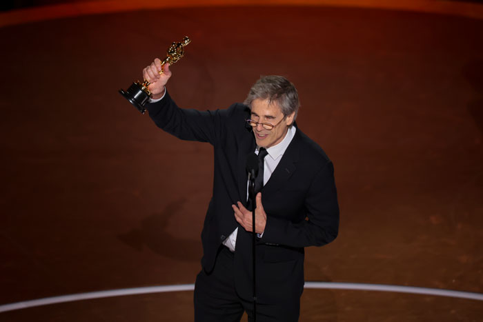 Man in a suit holding an Oscar, speaking at the Oscars 2025 ceremony stage. Man in a suit holding an Oscar, speaking at the Oscars 2025 ceremony stage.