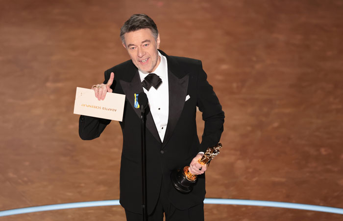 Man in tuxedo holding Oscar trophy on stage at the 2025 Oscars ceremony. Man in tuxedo holding Oscar trophy on stage at the 2025 Oscars ceremony.