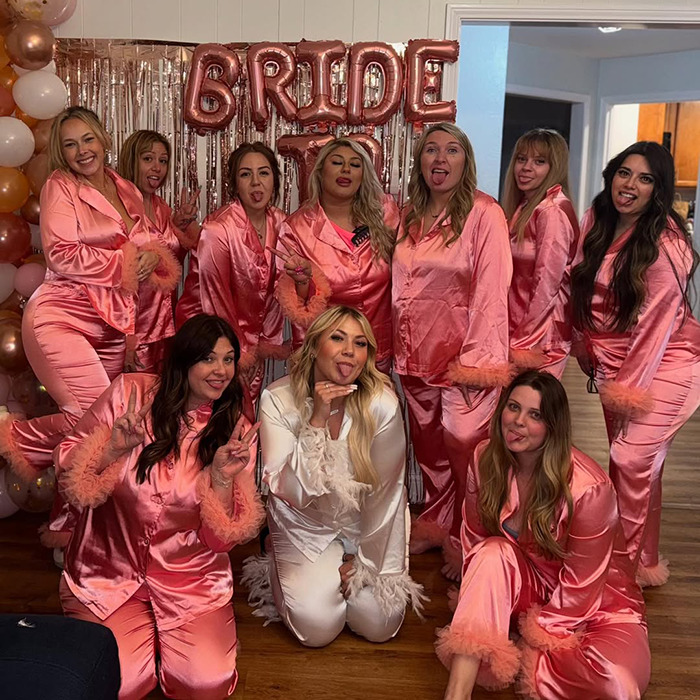 Bride-to-be in white poses with friends in pink pajamas at her bachelorette party, under "bride" balloons. Bride-to-be in white poses with friends in pink pajamas at her bachelorette party, under "bride" balloons.