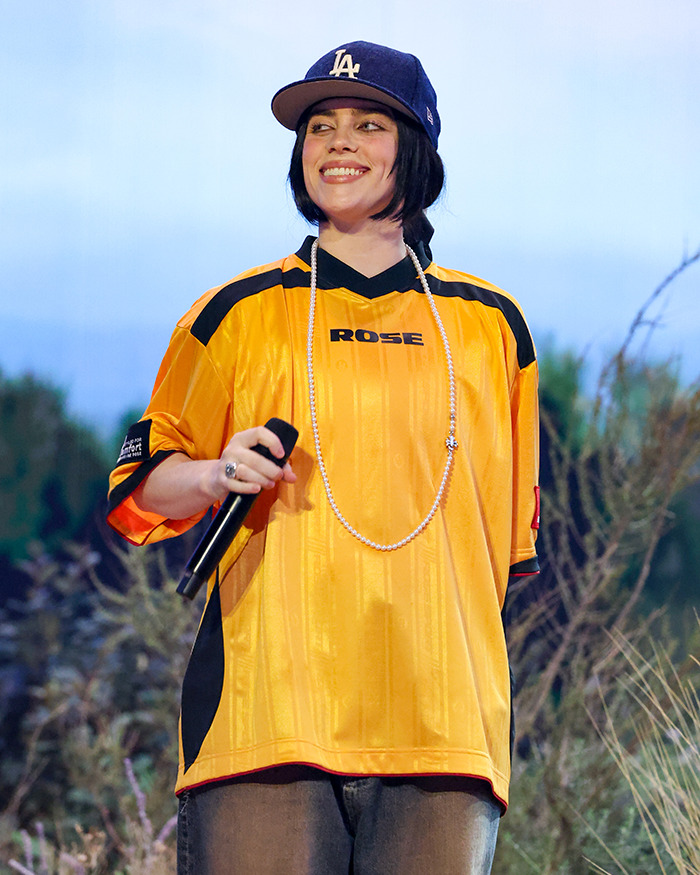 A person resembling Billie Eilish in a yellow jersey and cap, holding a microphone outdoors. A person resembling Billie Eilish in a yellow jersey and cap, holding a microphone outdoors.