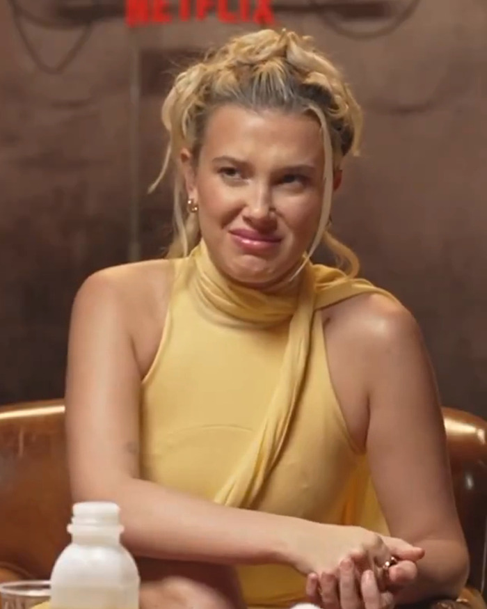 Woman in a yellow outfit sitting, with a Netflix backdrop, making an expressive face during an interview. Woman in a yellow outfit sitting, with a Netflix backdrop, making an expressive face during an interview.