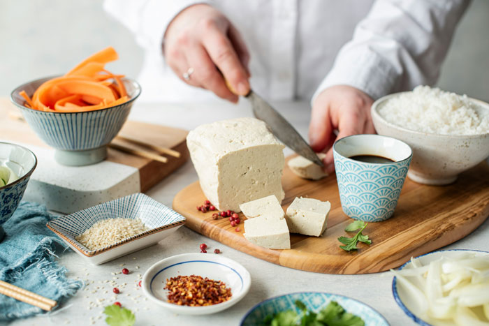 Person slicing tofu on a wooden board, surrounded by vegetables, a bowl of rice, and various spices, related to cooking. Person slicing tofu on a wooden board, surrounded by vegetables, a bowl of rice, and various spices, related to cooking.