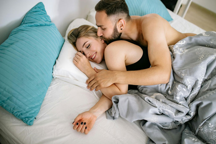Couple cuddling in bed, both smiling, with gray blankets and blue pillows, highlighting open relationship dynamics. Couple cuddling in bed, both smiling, with gray blankets and blue pillows, highlighting open relationship dynamics.