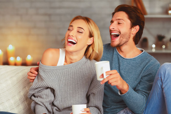 A couple laughing on a sofa, each holding a mug, in a cozy living room setting. A couple laughing on a sofa, each holding a mug, in a cozy living room setting.