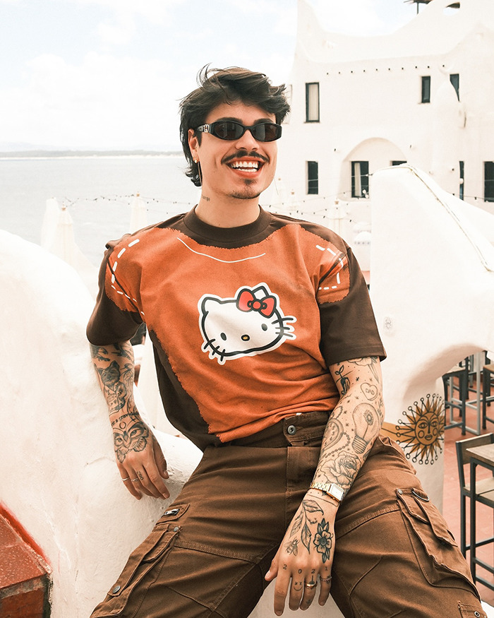 Tattooed person in sunglasses and Hello Kitty shirt, sitting outdoors with Brazilian architecture in background. Tattooed person in sunglasses and Hello Kitty shirt, sitting outdoors with Brazilian architecture in background.