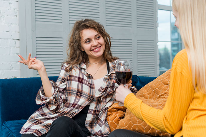 Two friends chatting on a couch, one holding a glass of wine, discussing wedding engagement plans. Two friends chatting on a couch, one holding a glass of wine, discussing wedding engagement plans.