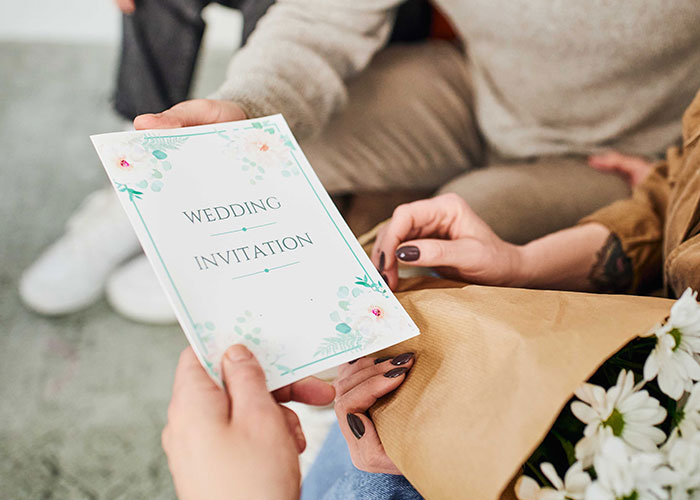 People holding a wedding invitation card, surrounded by flowers and casual attire, suggesting a discussion about wedding plans. People holding a wedding invitation card, surrounded by flowers and casual attire, suggesting a discussion about wedding plans.