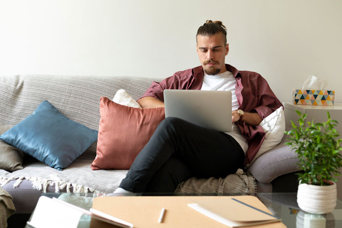 Man sitting on sofa, using laptop, considering moving in with brother after losing house, with plants and cushions nearby. Man sitting on sofa, using laptop, considering moving in with brother after losing house, with plants and cushions nearby.