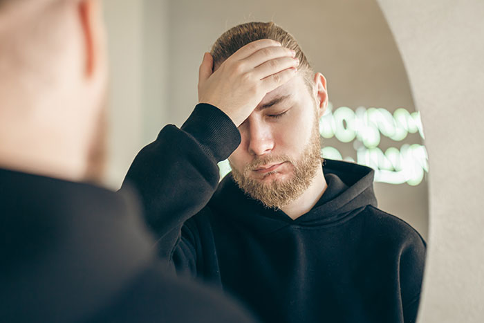 Man in black hoodie looking distressed, hand on forehead, in front of a mirror. Man in black hoodie looking distressed, hand on forehead, in front of a mirror.