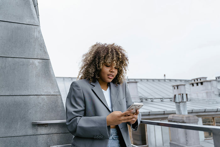 Woman standing outside in a gray blazer, looking at her phone with concern, representing permission issues. Woman standing outside in a gray blazer, looking at her phone with concern, representing permission issues.