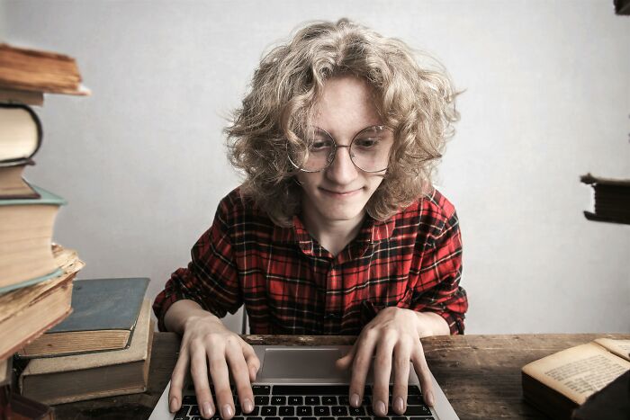 Person with curly hair and glasses typing on a laptop, surrounded by books, highlighting ridiculous bullying topics.