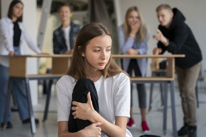 A girl sits alone, looking sad, while peers in the background appear to mock, illustrating bullying.