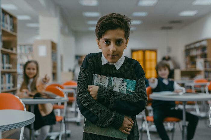 A boy holding books, standing in a classroom while two other children point fingers at him, illustrating bullying over ridiculous things.