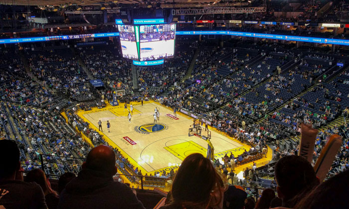 Fans watch a basketball game in a large arena with a prominent scoreboard displaying the action. Fans watch a basketball game in a large arena with a prominent scoreboard displaying the action.