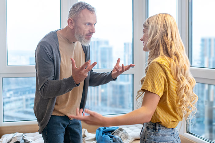 Stepdad arguing with a 27-year-old woman at home, both using expressive gestures near a large window. Stepdad arguing with a 27-year-old woman at home, both using expressive gestures near a large window.