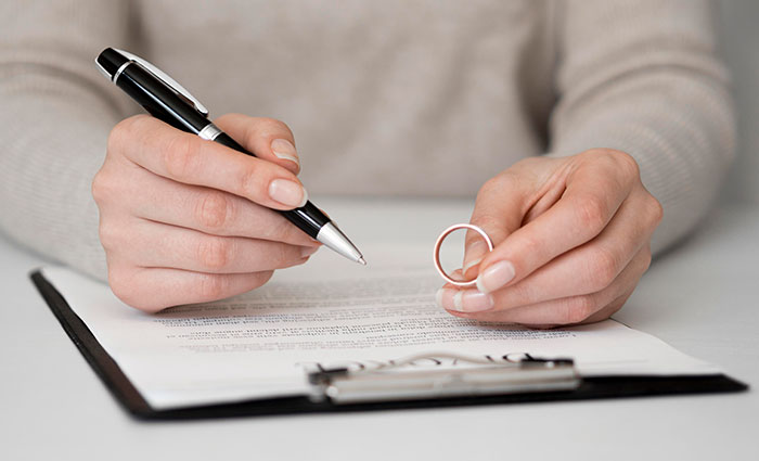 Person holding a wedding ring and pen over divorce papers, symbolizing affair partners' emotional conflict. Person holding a wedding ring and pen over divorce papers, symbolizing affair partners' emotional conflict.