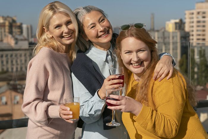 Three smiling women enjoying drinks on a rooftop, representing a childfree friend group gathering. Three smiling women enjoying drinks on a rooftop, representing a childfree friend group gathering.