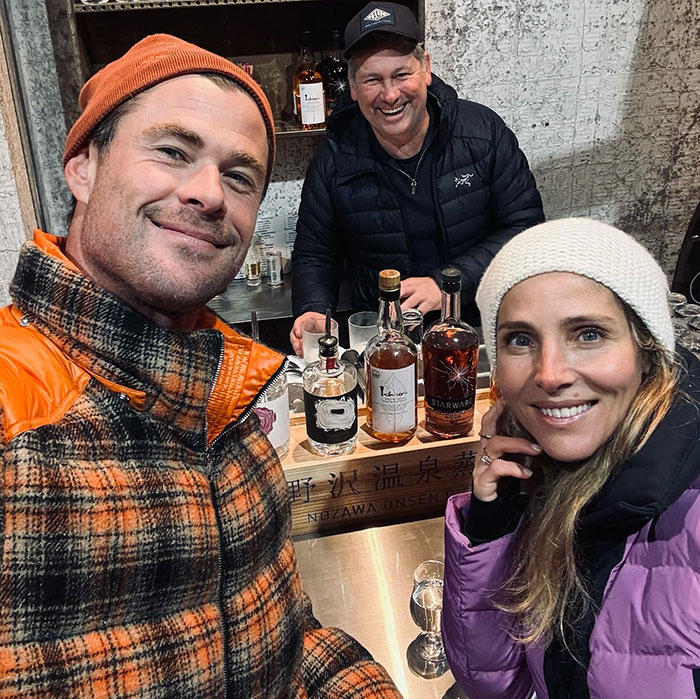 Two people smiling in front of a bar, wearing winter clothing, bottles visible on the counter. Two people smiling in front of a bar, wearing winter clothing, bottles visible on the counter.
