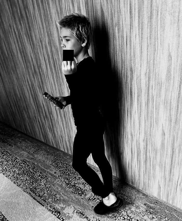 Young boy in black attire holding a remote, middle finger censored, leaning against a wooden wall. Young boy in black attire holding a remote, middle finger censored, leaning against a wooden wall.