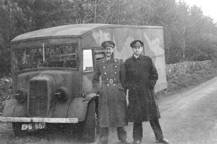 Two men in military coats stand in front of a vintage truck on a rural road, reminiscent of historical events.