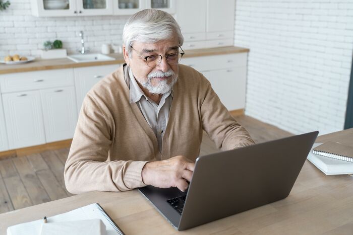 Elderly man with glasses typing on a laptop at a kitchen table, embodying weirdest things society accepts as normal.
