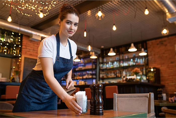 Young waitress in a cafe setting up a table, wearing a white shirt and apron, under warm lighting. Young waitress in a cafe setting up a table, wearing a white shirt and apron, under warm lighting.