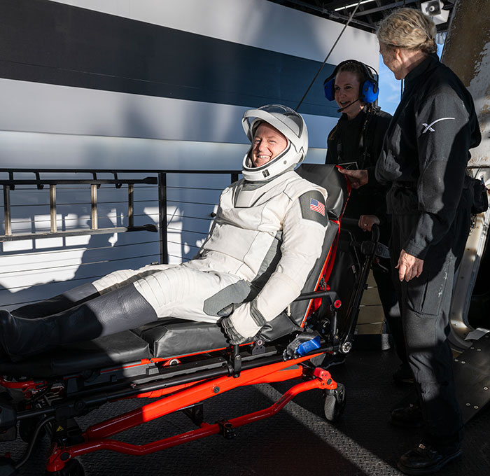 NASA astronaut returns to Earth, smiling in space suit, attended by crew on a stretcher. NASA astronaut returns to Earth, smiling in space suit, attended by crew on a stretcher.