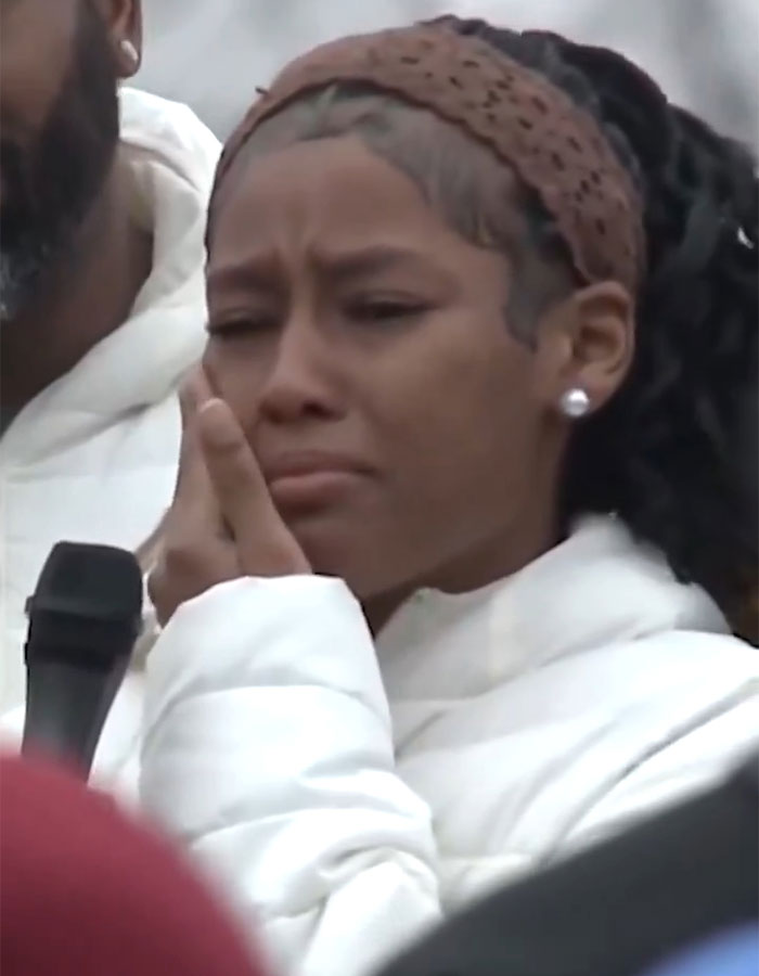 Track star in white coat, emotional at support rally, holding a microphone. Track star in white coat, emotional at support rally, holding a microphone.