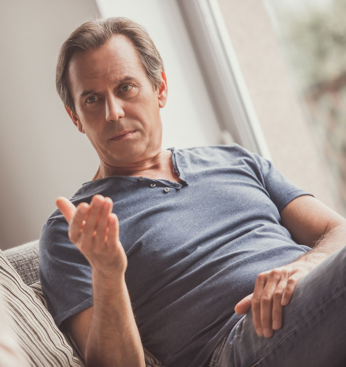 Man in a blue shirt seated on a couch, gesturing with his hand, discussing family care responsibilities. Man in a blue shirt seated on a couch, gesturing with his hand, discussing family care responsibilities.