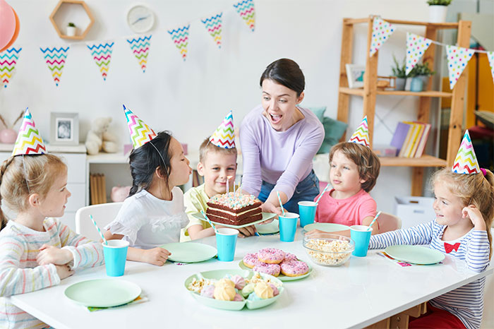 Children celebrate a birthday party with colorful hats, cake, and snacks, while a mom brings a chocolate cake to the table. Children celebrate a birthday party with colorful hats, cake, and snacks, while a mom brings a chocolate cake to the table.
