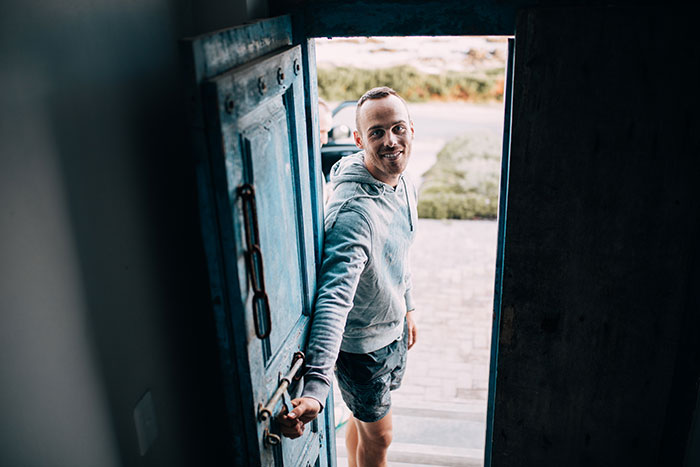 A man smiling while opening a blue door, wearing a gray hoodie and shorts, resembling a brand ambassador.