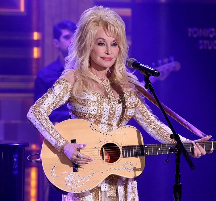 Dolly Parton performing on stage, holding a guitar, in a sparkling outfit. Dolly Parton performing on stage, holding a guitar, in a sparkling outfit.