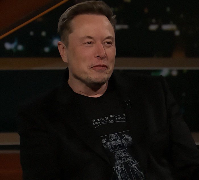 A person in a black suit appears on a talk show set. A person in a black suit appears on a talk show set.