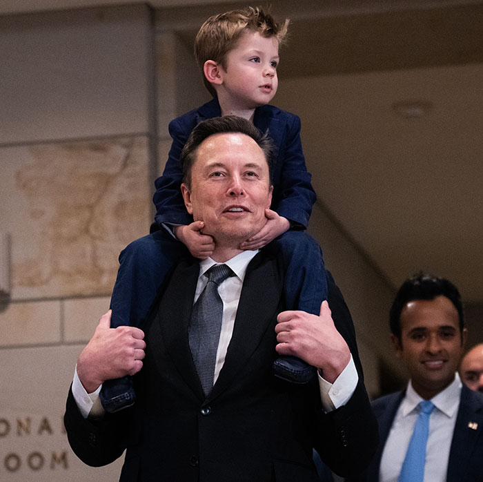 Man in a suit carrying a child on his shoulders indoors, with people in the background. Man in a suit carrying a child on his shoulders indoors, with people in the background.