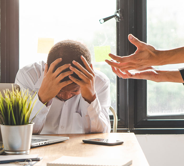 Man in distress at work desk, head in hands, with another's hands gesturing, representing stress from a toxic boss. Man in distress at work desk, head in hands, with another's hands gesturing, representing stress from a toxic boss.